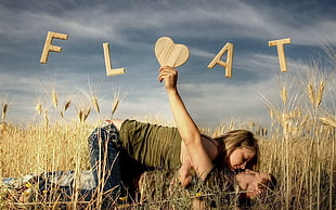 woman in green tank top over man in black t-shirt on grass field holding beige wooden heart decor forming float signage