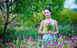 selective focus photography of woman wearing pink and blue strap dress holding purple petaled flowers
