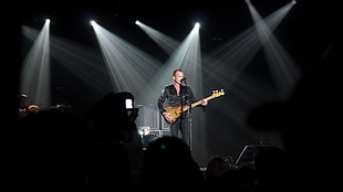 man in black jacket and black pants outfit playing brown guitar and singing standing on stage