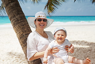 photo of woman holding baby leaning on coconut tree on white sand beach during day time