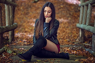 shallow focus photography of woman in black leather jacket sitting on brown wooden dock during daytime