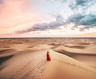 woman in red long dress walking in desert under cloudy sky during daytime
