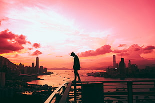 silhouette of standing person wearing fitted pants and cap on railings with distance at high rise buildings across body of water under blue sky
