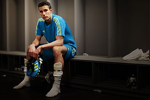 man in blue Adidas jersey shirt holding blue cleats sitting on bench