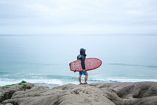 man in black hoodie carrying red surfboard standing on rock formation