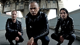 three men wearing black leather jackets squatting on gray concrete pavement