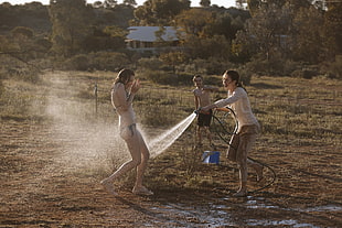 woman in black bikini getting wet by woman in beige long-sleeved shirt holding black garden hose