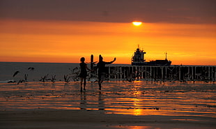 silhouette photo of two person running on brown sand in front of body of water during sunset day