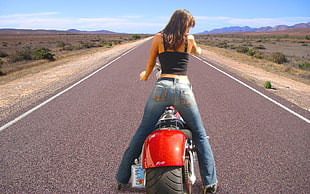 woman in black thick-strap shirt and blue jeans riding red cruiser motorcycle in the middle of gray asphalt road during daytime