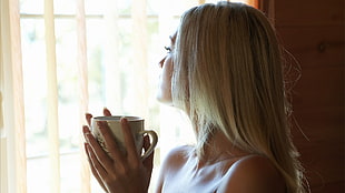 woman holding white ceramic teacup while looking outside the window