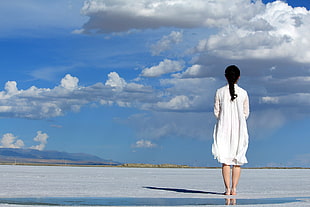 woman in white long sleeved dress standing on sand under blue sky and white cumulus clouds
