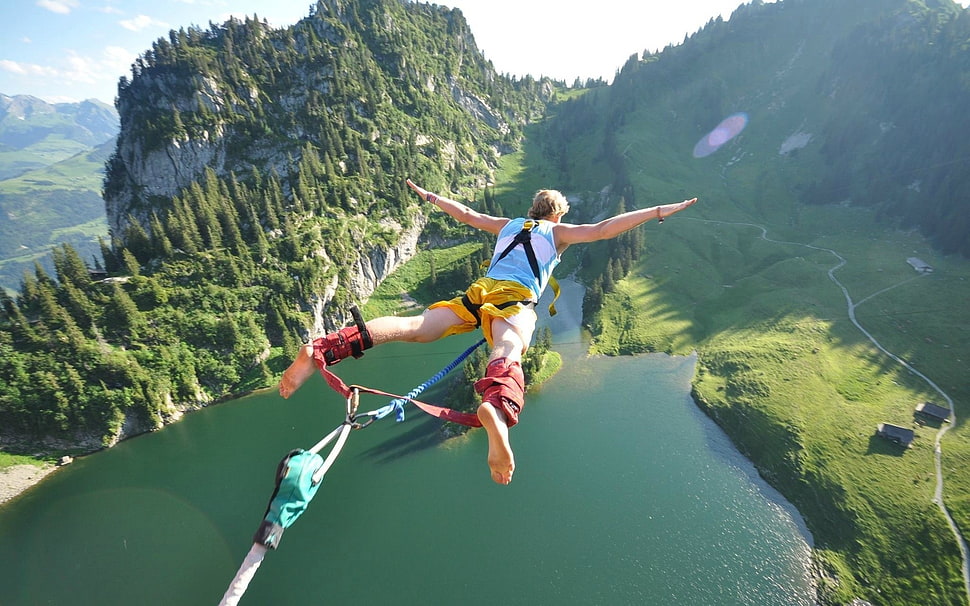 man in blue tank top jumped over body of water near pine trees during daytime HD wallpaper