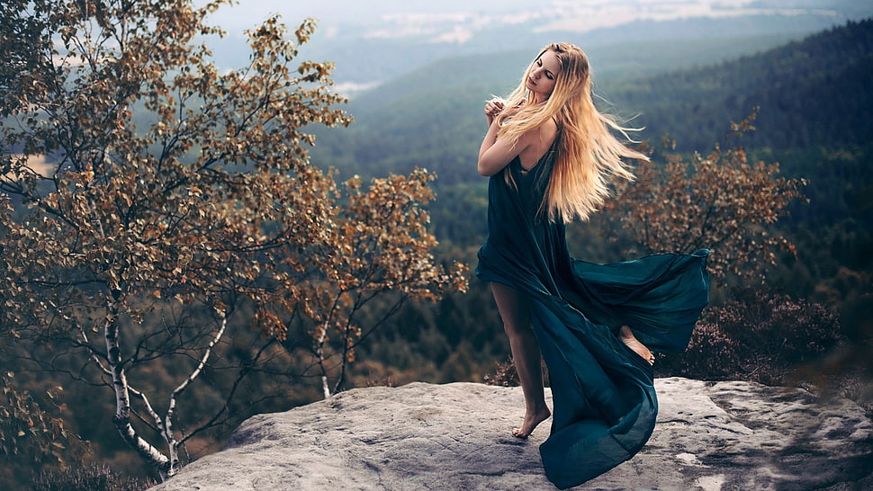 woman wearing teal sleeveless dress posing with both of her hands on her heart while she close her eyes on grey cliff surrounded with brown trees HD wallpaper