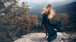 woman wearing teal sleeveless dress posing with both of her hands on her heart while she close her eyes on grey cliff surrounded with brown trees