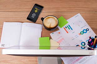 mug filled with latte infront of a computer monitor beside an open notepad