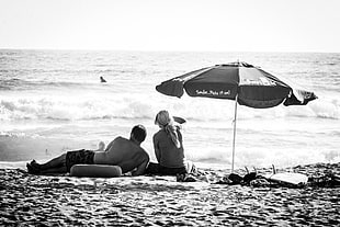 grayscale photo of couple sitting under umbrella near body of water