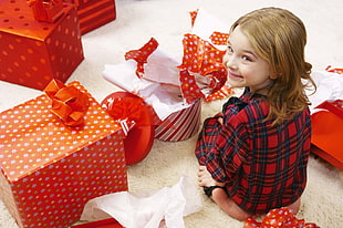 girl in red and green plaid pajama set sitting in front of wrapped and opened Christmas gift boxes