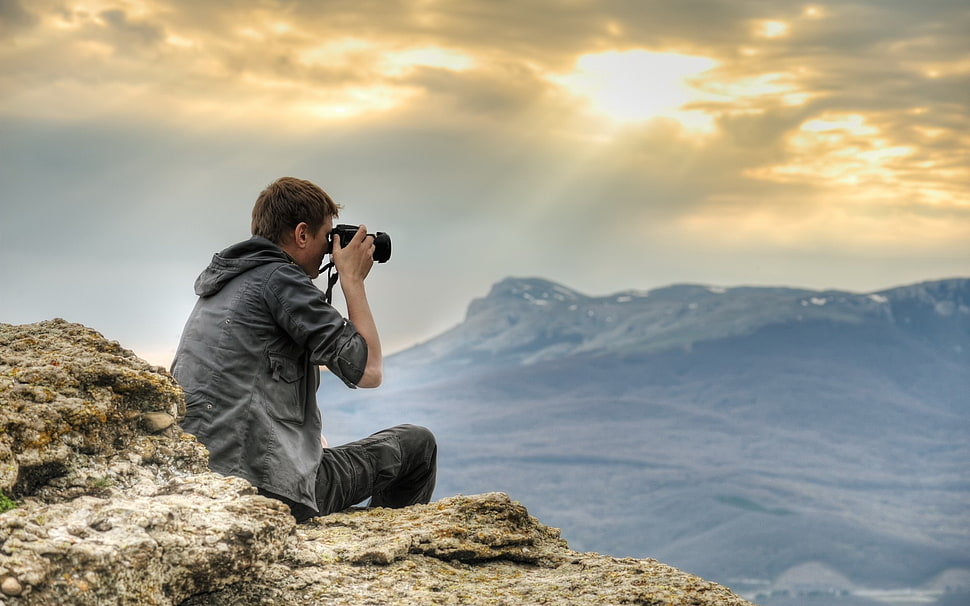 man in black denim hoodie holding DSLR camera sitting on rock during daytime HD wallpaper