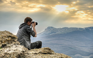 man in black denim hoodie holding DSLR camera sitting on rock during daytime