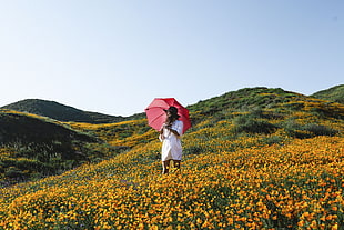 woman in white midi dress holding red umbrella taking picture on yellow flower field on during daytime