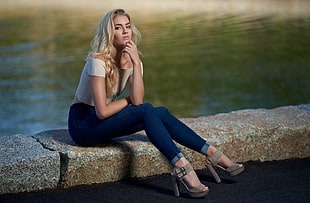 woman wearing white off-shoulder shirt, blue jeans and brown peep-toe platform pumps sitting on brown concrete pavement near body of water at daytime