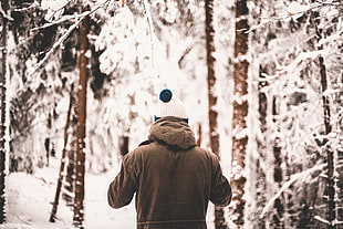 person wearing hooded jacket and white bobble knit cap standing near the trees covered with snow