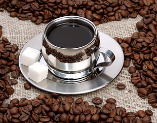 gray steel coffee cup filled with brown liquid on top of saucer with two white sugar cubes and coffee beans