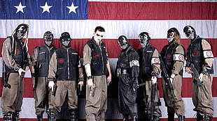 eight men in gray boiler suits and black leather vest holding guns with U.S.A flag background