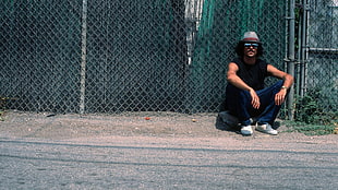 sitting man wearing gray-and-brown fedora hat, black tank top, and blue denim jeans behind wire fence over concrete ground