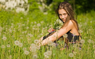 woman holding bunch of flowers sitting on flower field wearing black dress