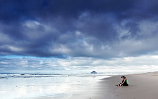 man wearing green top sitting facing on seashore during nimbus clouds