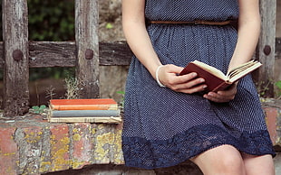 woman wearing blue mini dress sitting on concrete surface reading book during daytime