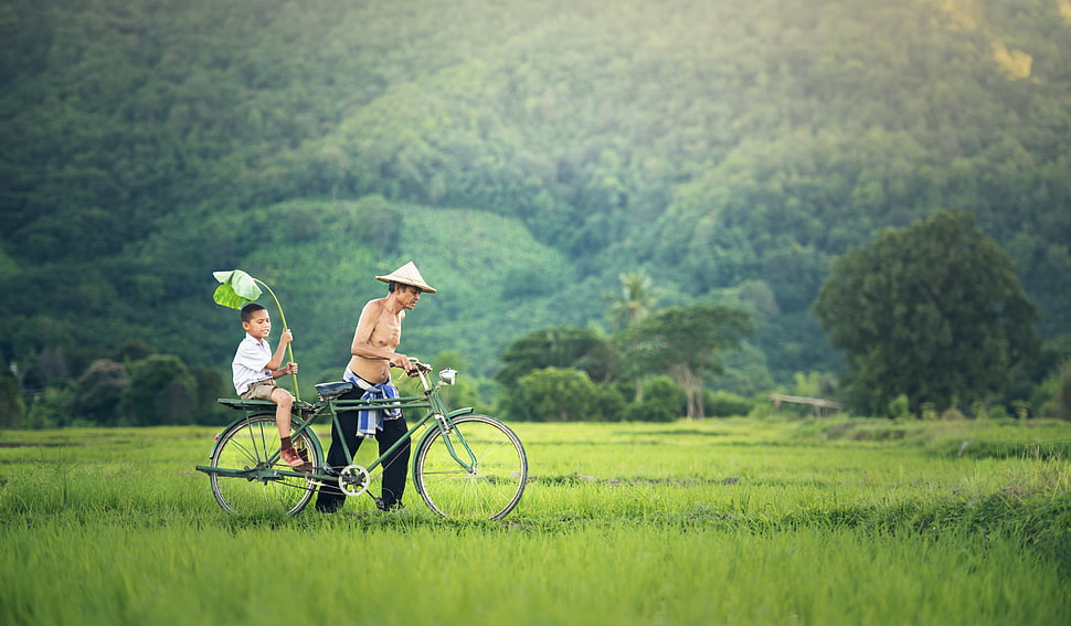 man wearing black pants and round hat pushing the green commuter bike with boy on green grass field near the mountain during daytime HD wallpaper