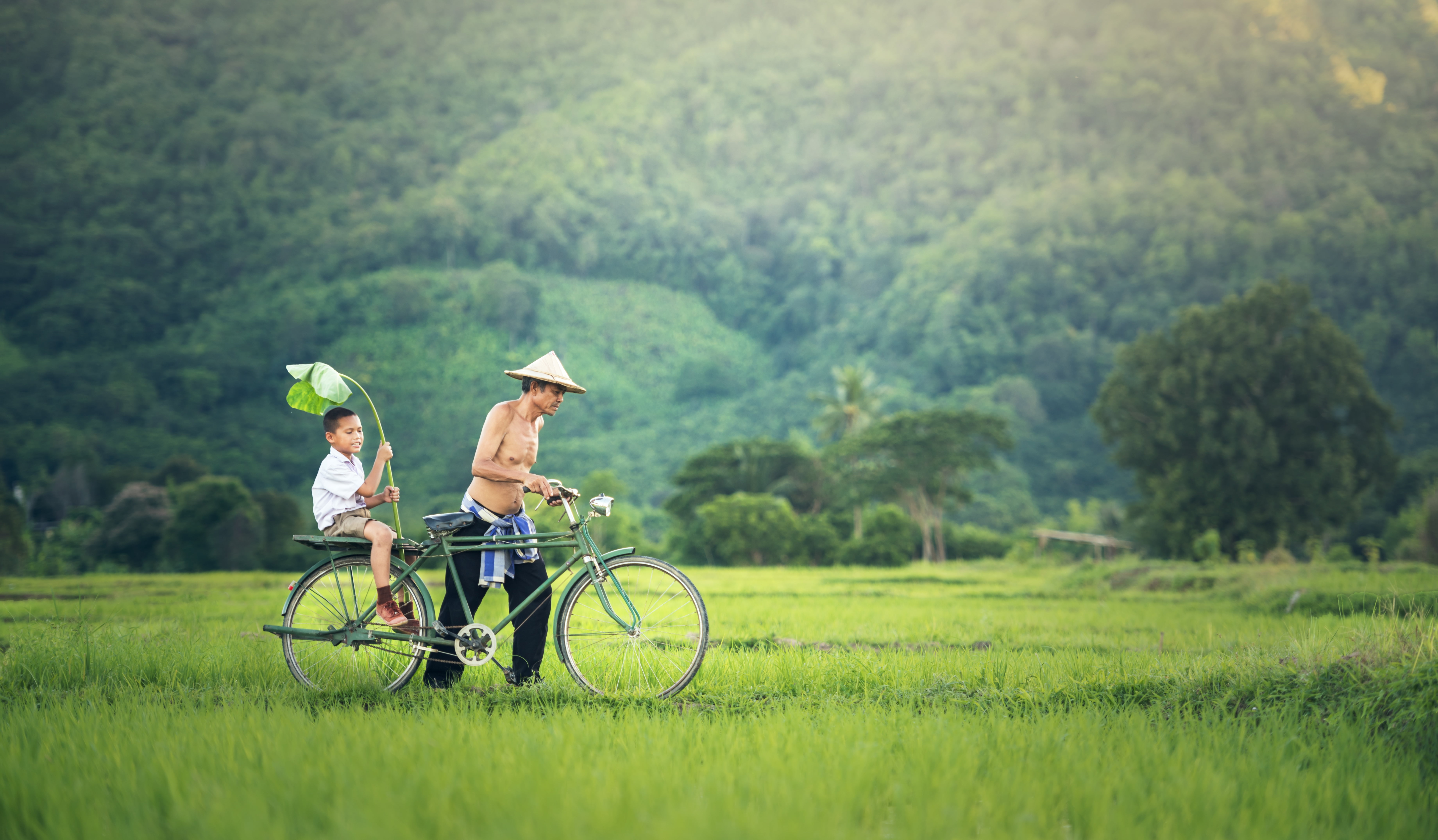 man wearing black pants and round hat pushing the green commuter bike with boy on green grass field near the mountain during daytime