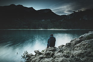 man sitting on rock in front of body of water during day time photo HD wallpaper