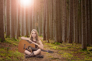 woman holding brown classical guitar sitting in the middle of the forest