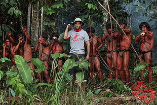 man in gray shirt together with native tribe holding handmade weapons aiming at camera inside deep forest during daytime