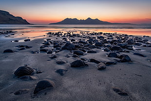 focus photo of grey stones on shore with a view of mountains afar at sunset HD wallpaper